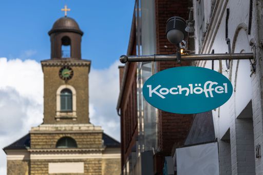 A hanging German kochloffel restaurant sign near a historic tower in Husum, Germany
