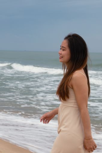 A smiling Asian woman walking on the sand across the water and enjoying the calming wave sounds