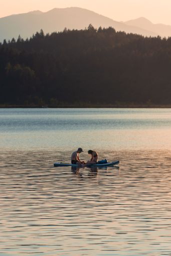 Two people sitting on a Stand-Up-Paddle in the middle of a scenic lake and enjoying the warm sunset