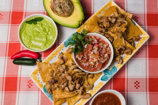 A top view of a plate of beef nachos next to dips and avocado, on a red checkered tablecloth