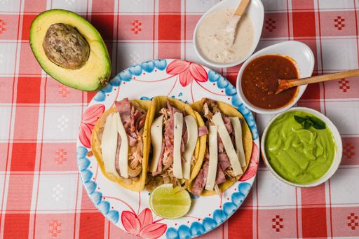 A top view of a plate of tacos next to dips and avocado, on a red checkered tablecloth
