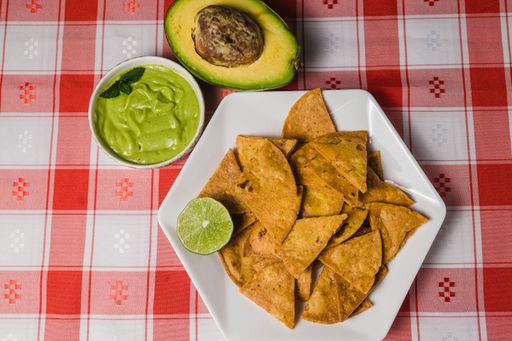 A top view of a plate of nachos next to dips and avocado, on a red checkered tablecloth