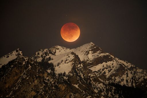 An orange Full Moon shining bright above a snow-capped rocky mountain at night