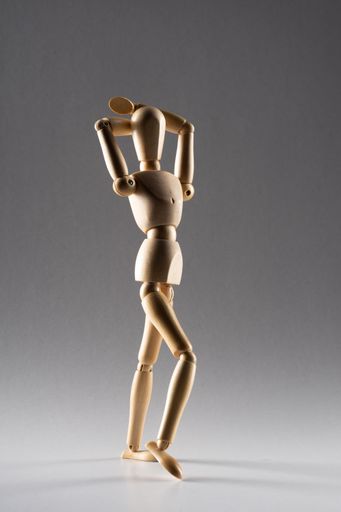A vertical shot of a wooden pose doll with both hands on its head standing in the shade