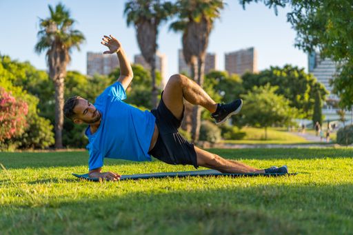 Hispanic man dressed in blue sportswear and mat doing a side plank in the park garden during sunset light.