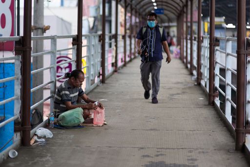 A closeup shot of a homeless man sitting on a bridge and another man with a face mask walking on it