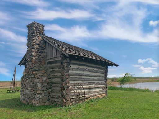 A stone-wooden small building on lush green lakeshore under blue cloudy sky