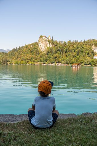 A vertical shot of a little boy sitting on the shore and looking at lake with Bled castle. Slovenia.