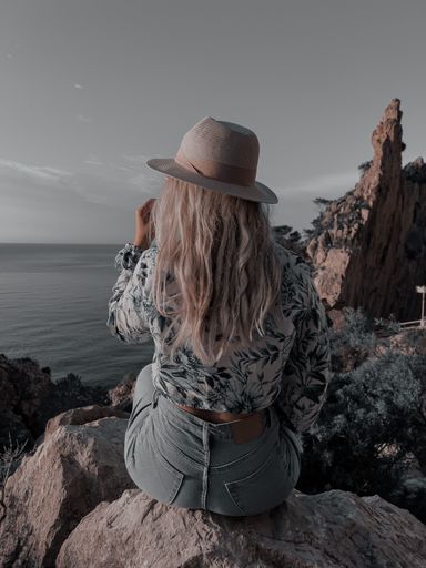 Vertical shot of young lady in hat sitting on rock enjoying the view of the calm sea and steep cliffs at sunset
