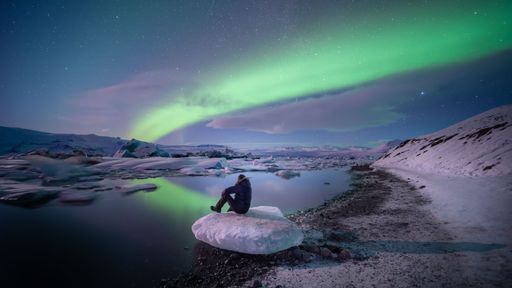 A breathtaking view of a male sitting on the stone in the lake on aurora night