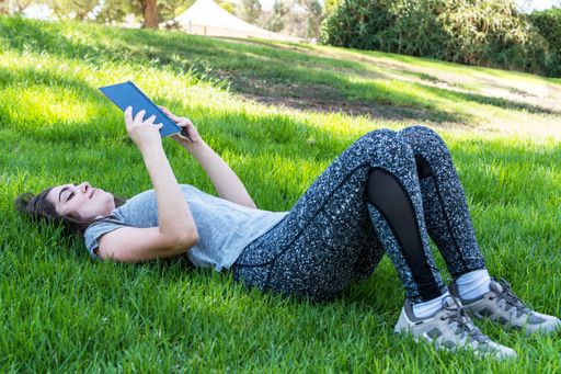 A closeup shot of a young Caucasian female reading a book on a tablet lying in a park