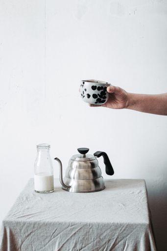 A vertical shot of a person holding a teacup over a teapot and a glass of milk