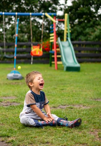 Young Polish Caucasian three years old boy sitting on green grass and cries close by playground equipment of a vacation resort on a summer day.