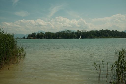 A gorgeous view of the Chiemsee with a sailing boat going across it, surrounded by lush greenery under a bright cloudy sky