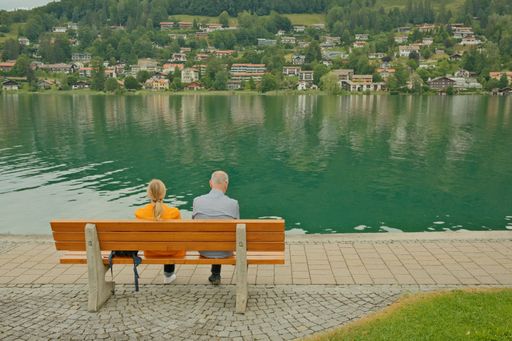 A man and a woman sitting together on a bench on the shore of the Tegernsee in Rottach-Egern, admiring the gorgeous view