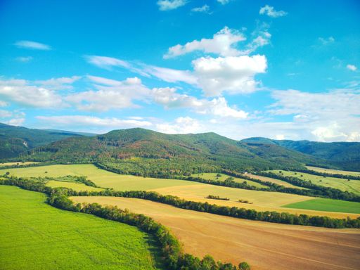 An aerial view of large fields and mountain forests under blue cloudy sky