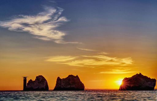 The Sun setting behind one of the rock stacks at The Needles Lighthouse on the Isle of Wight. The sky shows a few light summer clouds.