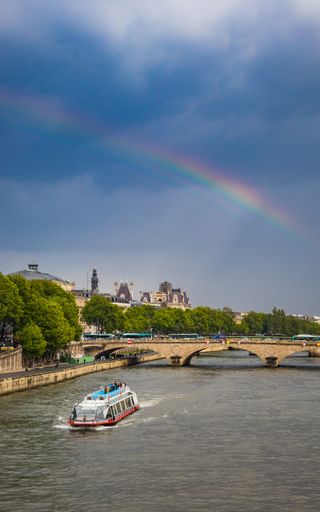 A vertical shot of boat floating on lake towards bridge under blue bright sky with rainbow