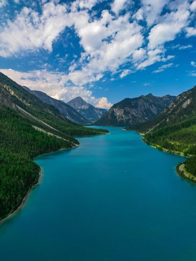 A gorgeous view of a bright blue river surrounded by tall mountains covered by vibrant green trees under a cloudy sky