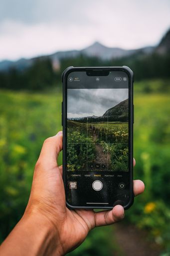 A man's hand holding a smartphone, taking a picture of a bright green field surrounded by mountains