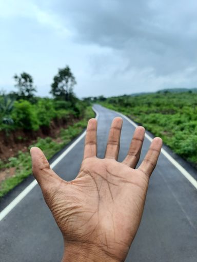 A vertical closeup of the palm of a hand with an empty road in between a green grass field on a cloudy day