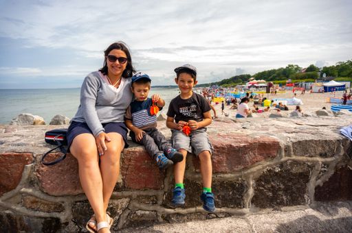Woman and two young boys sitting on a small wall in front of a crowded beach on a cloudy day.