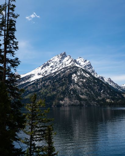 a vertical shot of snow capped mountain peak with lake in front of it during the winter in the sun.
