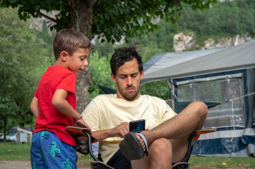 A Caucasian man using his phone, sitting in a chair at a campsite with his son