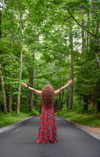 A vertical shot of a woman in a red dress standing on an empty road in a forest with happy open arms