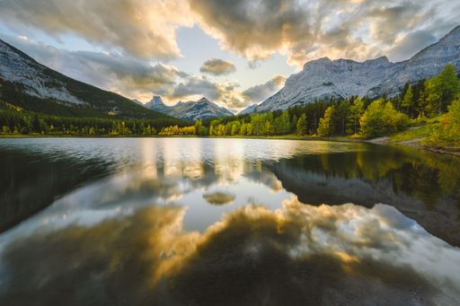 A beautiful view of a pond reflecting the green trees on the shore surrounded by snowy mountains under a cloudy sky