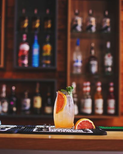A glass cup of Mezcal Grapefruit with slice on bar table with blur bottle shelves