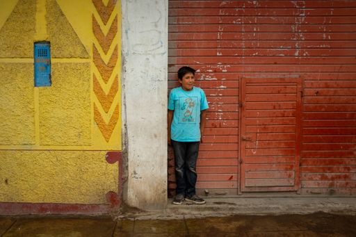 Ayacucho, Huamanga / Peru - January 6 2016: Young Latin Boy in Blue Shirt Hiding from the Rain Under a Small Portion of Roof in a Yellow and Red Wall