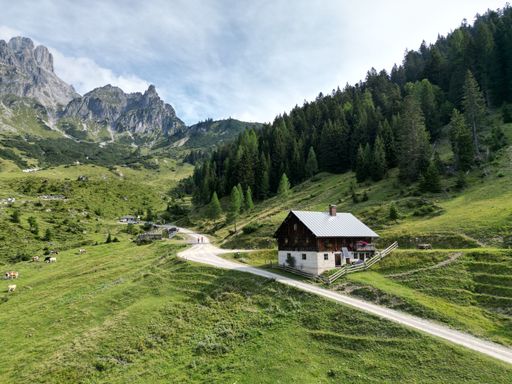 A beautiful view of an Alpine cottage next to the forest and mountains under a cloudy sky.