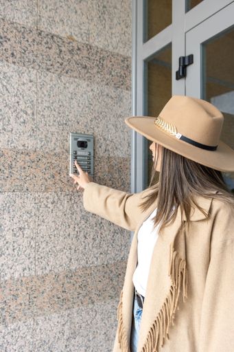 An adult female stands outside the doorway of a residential building, looking up and pressing the doorbell