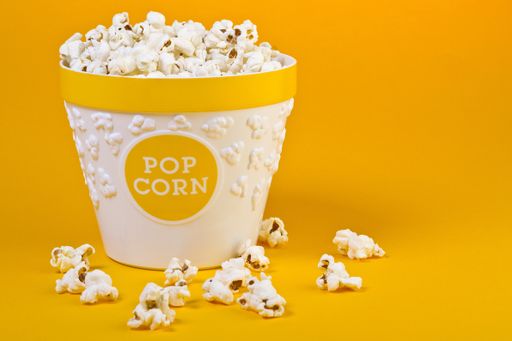 A white basket full of popcorn and some popcorn around it on an orange background
