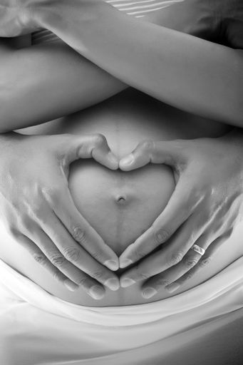 A vertical gray scale shot of two hands shaped like a heart on a pregnant mother's stomach