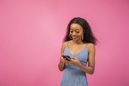 A closeup shot of a pretty Afro-American girl in an exciting mood holding her phone on a pink background