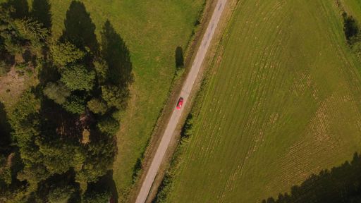 An aerial top view of a single red car driving on a highway surrounded by fields