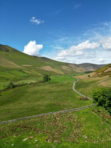 A vertical shot of a narrrow road through a green field and hills on a sunny summer day