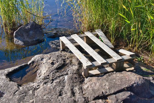 A closeup shot of an old wooden pallet on the shore, above the rock and a pond, with green grass in the background, on a sunny day