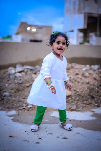 A portrait of a cute little girl smiling and standing on the ground in white dress and green trousers