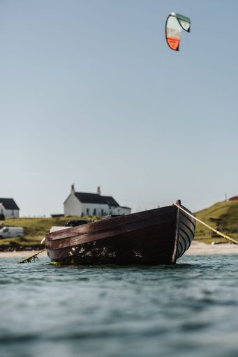 A selective focus on a small boat anchored on the sea, with houses on the Tiree's coast in the background, and a colorful kite flying above it