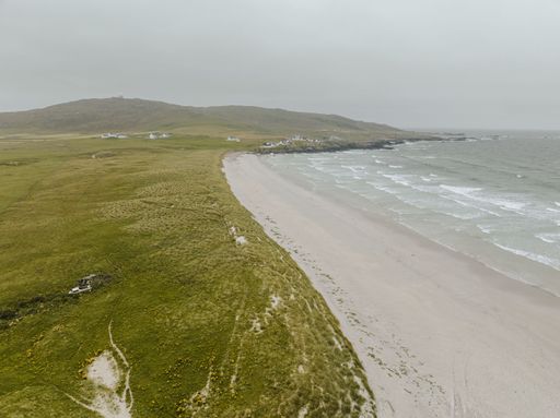 A beautiful view of a wavy sea and sandy beach of the Tiree island in Scotland, with a green field and hill in the background, on cloudy weather