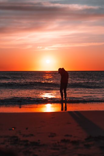 A vertical shot of a silhouette of a man standing on a beach during the sunset