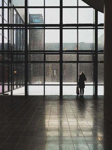 A vertical shot of a woman standing by a big window while snowing