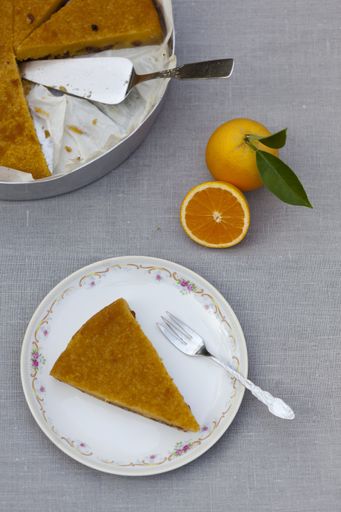 A slice of tasty orange pie in a white plate beside the whole pie and oranges on a gray surface