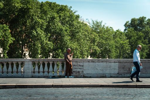 A closeup shot of two people walking on the sidewalk of a bridge with green trees in the background, Rome