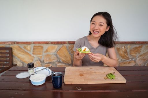 A vertical of an East Asian woman eating avocado toast she made.