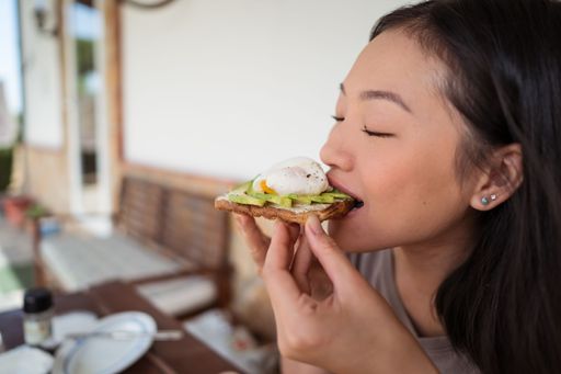 A vertical of an East Asian woman eating avocado toast she made.
