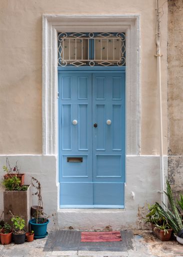 A vertical shot of a vintage blue front door of a beige building with plants at the entrance in Valetta, Malta